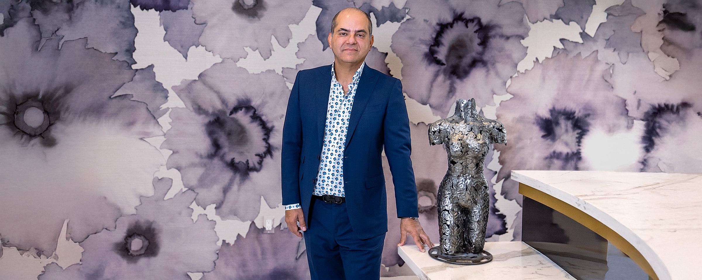 Dr Agha in suit with floral background and female body metal sculpture.